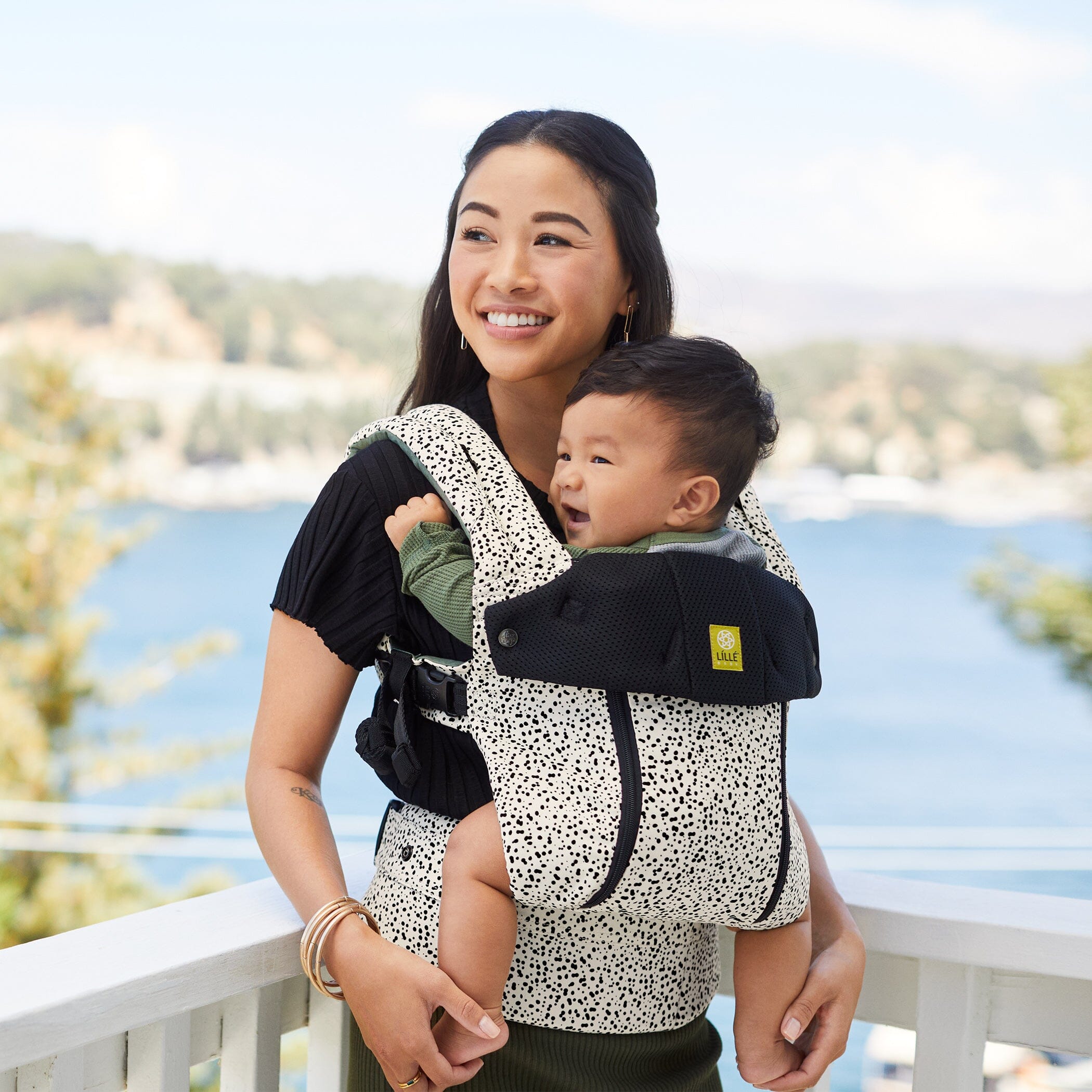 Mother standing on white balcony with child in baby carrier. A lake is behind them.