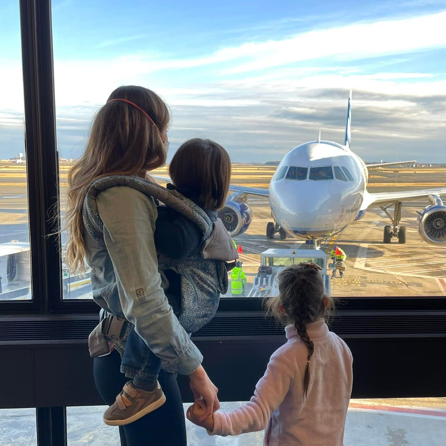 Woman holding a child's hand with another in a Lillebaby carrier looking out a window at an airplane