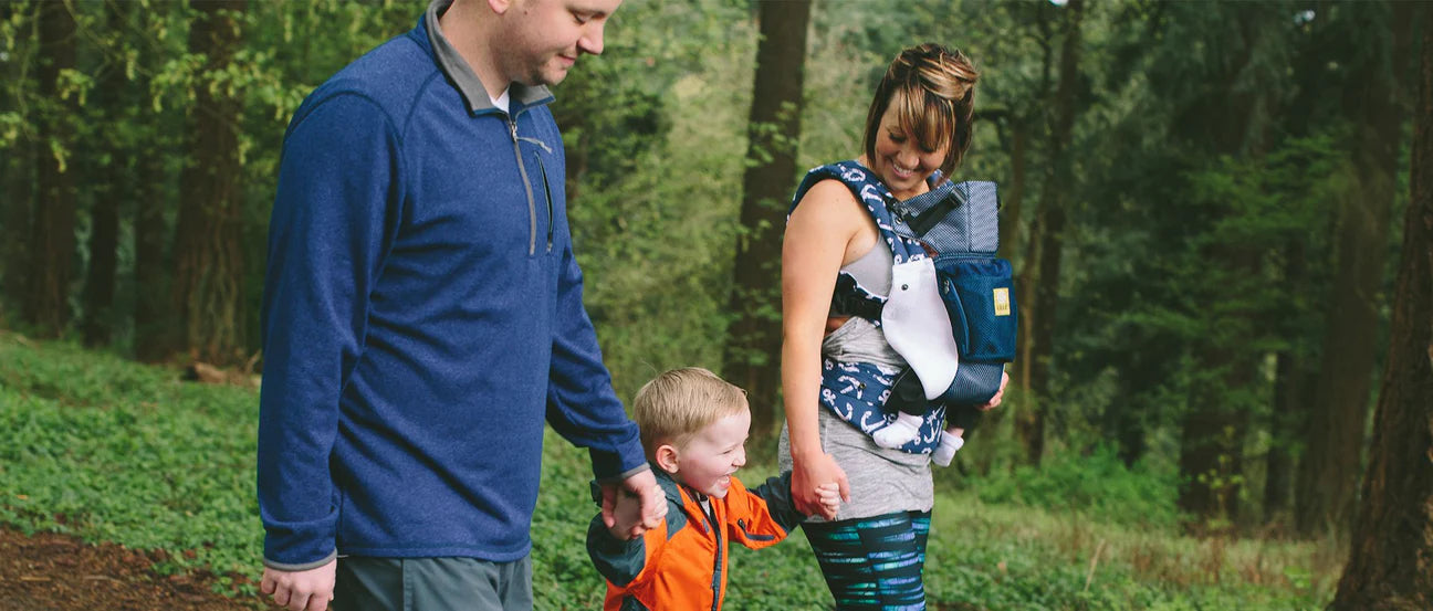Father and Mother walking through the woods, holding hands with son and carrying baby in lillebaby carrier