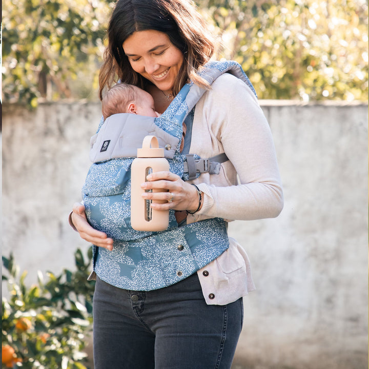 mom wearing baby in complete luxe in starfall while holding cream colored bink bottle