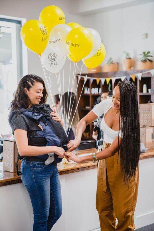 Mom ties balloons onto another mom's LILLEbaby carrier during a party.