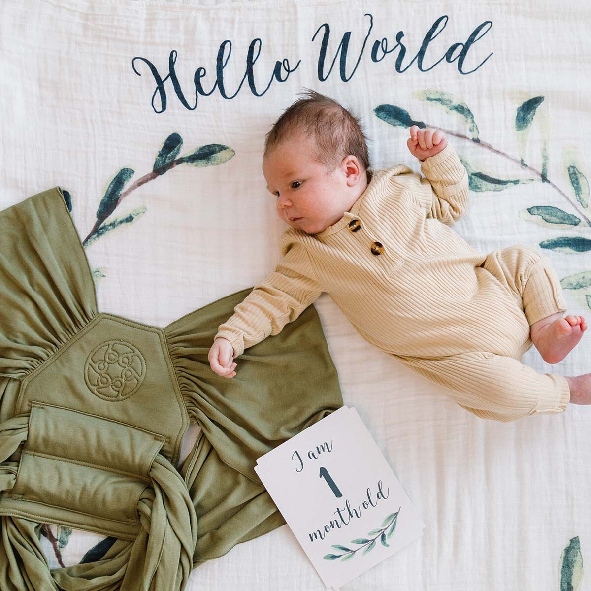 1 month old baby on lulujo blanket in hello world pattern with lillebaby's dragonfly wrap in moss next to baby